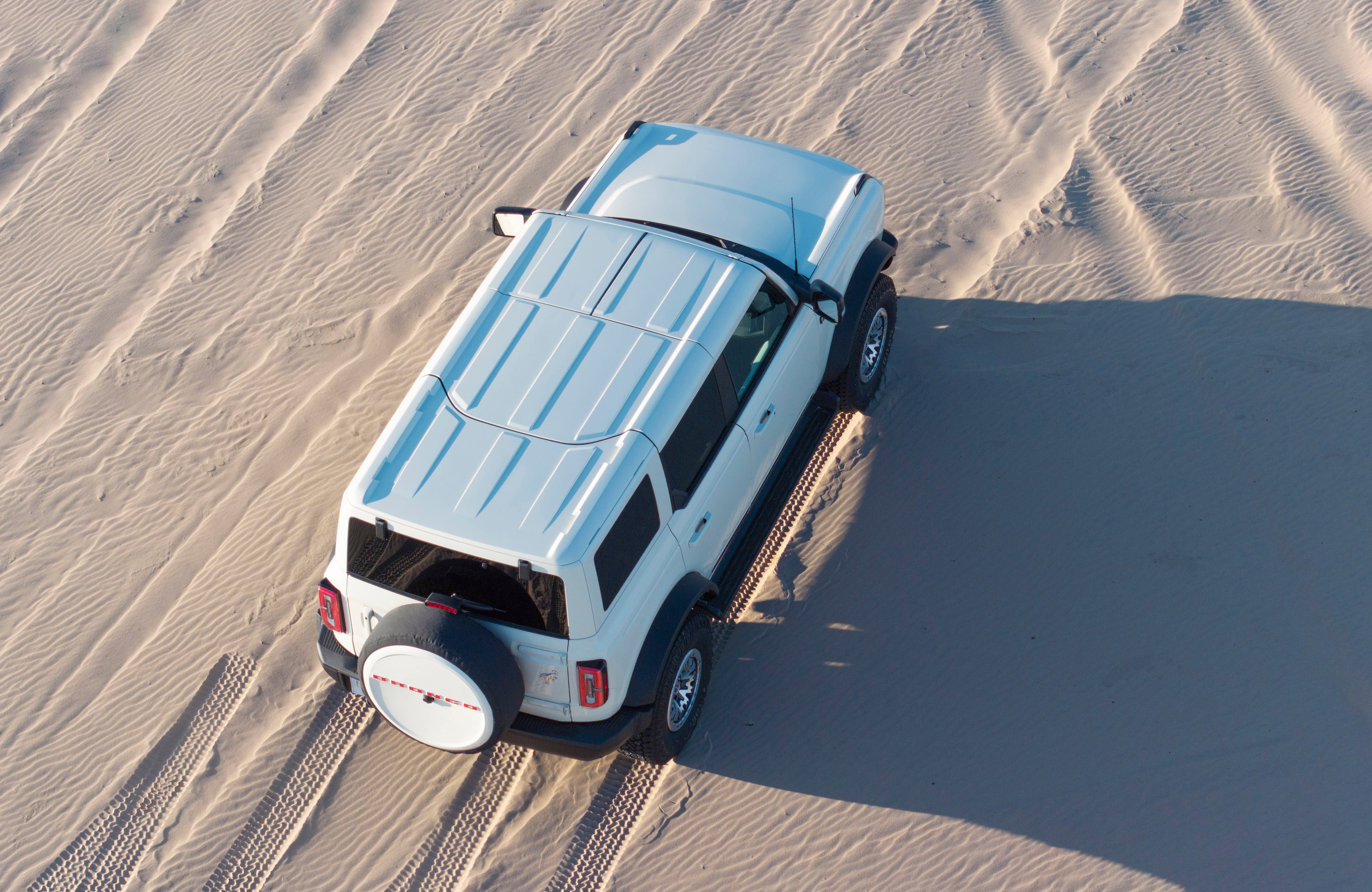 Over-head shot of a white 2026 Bronco 60th Anniversary Package vehicle. White with red accents. Vehicle pictured parked on sand. 